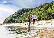 Spaziergang, Paar spaziert am Strand entlang, idyllische Küstenlandschaft., Paar spaziert am Strand entlang, umgeben von grünen Hügeln.